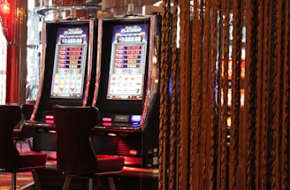 A pair of casino slot machines, each with detailed screens displaying various game outcomes and jackpot amounts. The chairs in front of the slot machines are dark and cushioned for comfort. The background features a hanging curtain made of golden ropes and other casino decorations are visible.