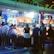 A group of people standing in front of an illuminated amusement arcade, engaging with various gaming machines. The environment is bustling with activity, and colorful lights create a vibrant atmosphere. The ceiling is decorated with large, playful images of dice and other game-related graphics, enhancing the lively mood.